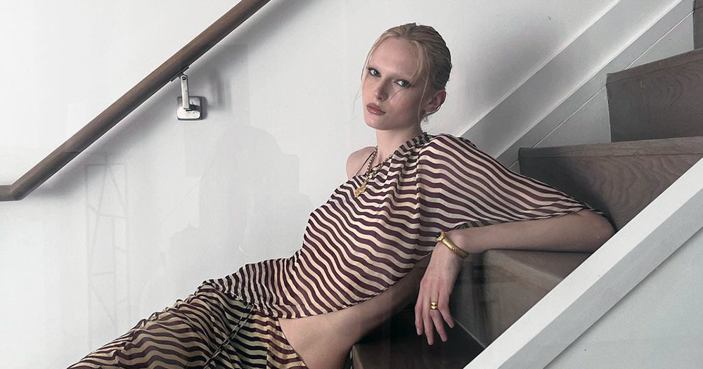 Alex Consani, who has been nominated for Model of the Year, laying on the stair while wearing a striped brown and white dress.