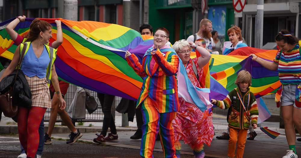 An image of Galway Pride from 2023. Several people march with a large Pride flag outdoors. One person wears a full rainbow suit.