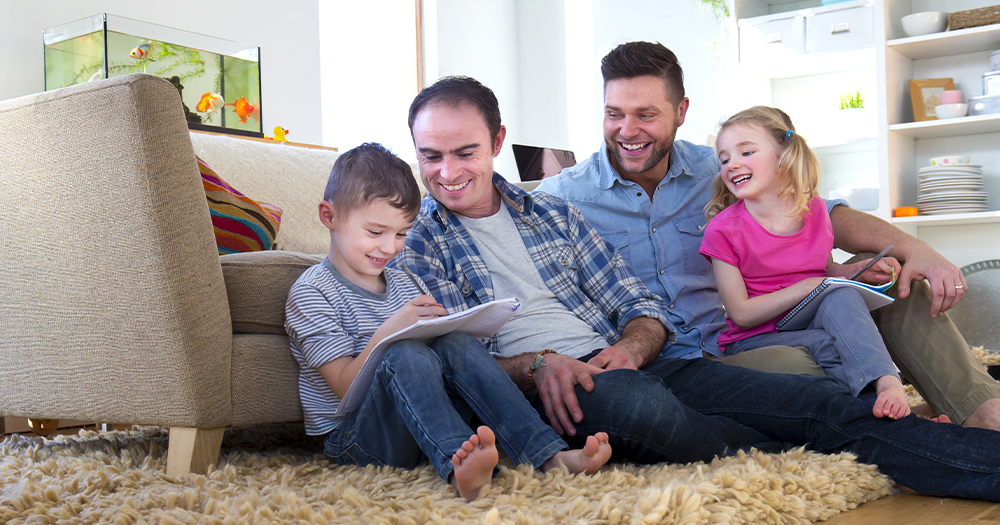 This article is about a surrogacy conference hosted by Irish Gay Dads. In the photo, two dads laughing and doing homework with their kids while sitting on the floor in front of a sofa.