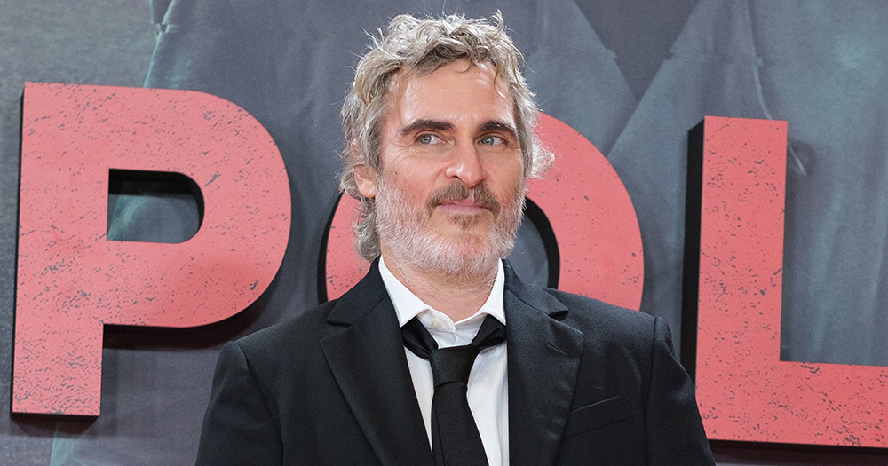 Joaquin Phoenix wearing a black suit, looking slightly to the side and smiling.