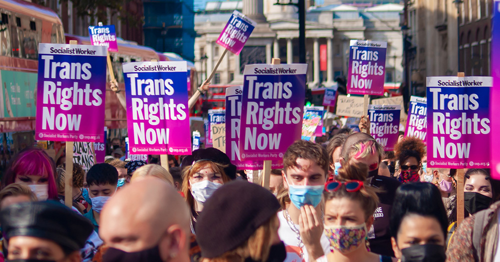 This article is about NHS England's plan to open six regional gender centres. The image shows people protesting in London holding signs that read "Trans Rights Now".