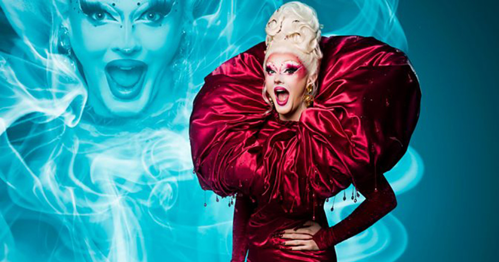 Belfast queen Charra Tea who will be competing on Drag Race UK. She wears an elaborate red dress and blonde wig and is photographed in front of a blue background.