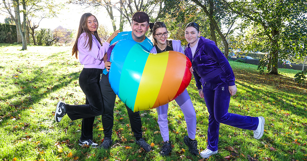 This article is about LGBTQ+ safety in school. The image shows four teenagers holding a rainbow balloon and smiling.