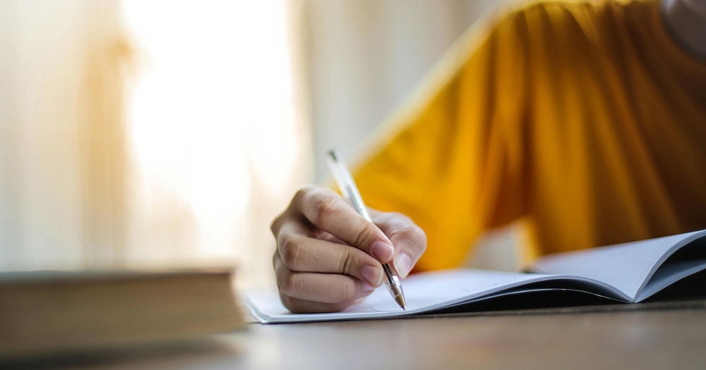 This article is about a new queer writing project in Ireland. The image shows a person in a yellow jumper writing with a pen and paper.