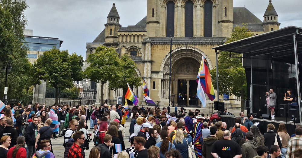 Photo from Trans Pride 2024 in Belfast, Ireland, with a crowd gathered in a square with a stage, waving rainbow flags.