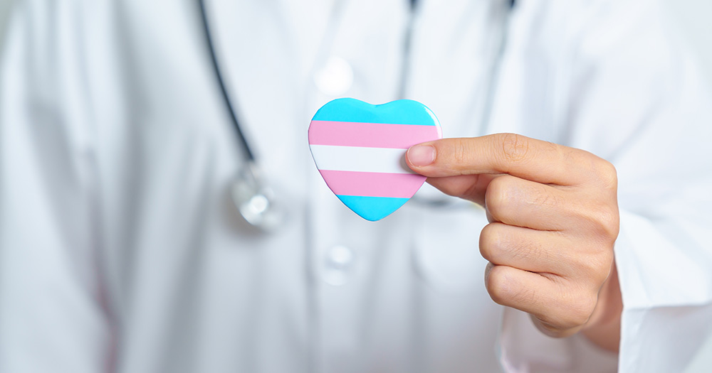 This article is about a new professional association for trans healthcare in Ireland. The image shows a person wearing a white medical cloak with a stethoscope around their neck holding a small loveheart in the colours of the trans flag.