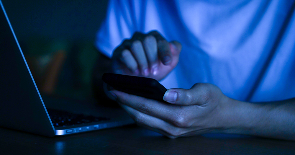 This article is about a Northern Irish man catfishing children. In the photo, the hands of a person holding a phone in the dark.