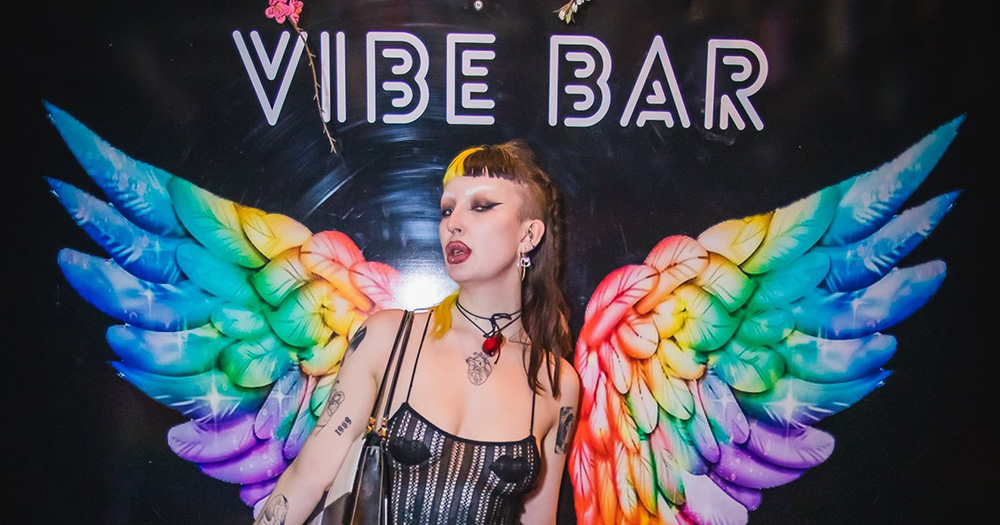 An image of a person standing in front of the Vibe Bar sign, with rainbow wings in the background.