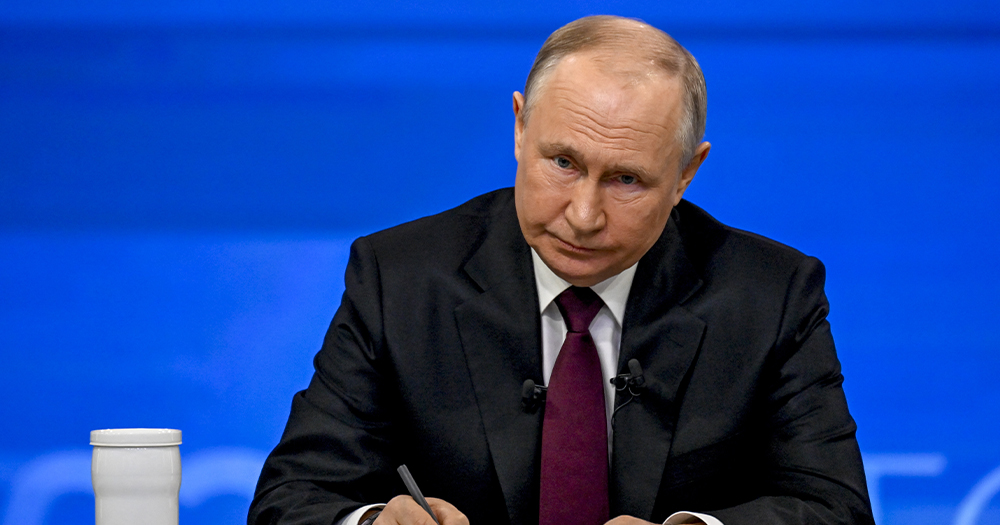 Photo of Russian President Putin, who recently signed a law banning adoptions for countries allowing gender-affirming care, sitting at a table and writing, with a blue background.