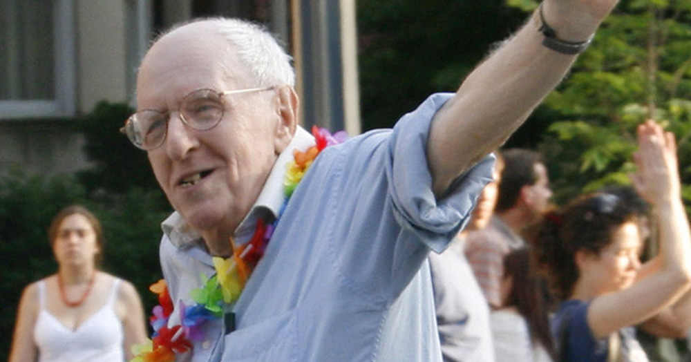 An image of Frank Kameny waving. He wears a rainbow floral necklace and a blue shirt.