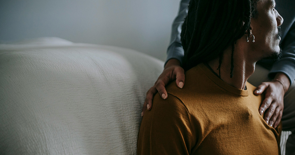 This article is about a new LGBTQ+ intimate partner abuse network. The image shows a person sitting against a bed on the right hand side, with another persons hands supportively on their shoulders.