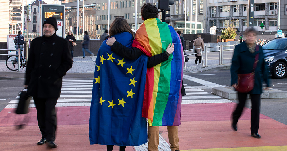 This article is about a petition calling for a conversion therapy ban. The image shows the backs of two people with their arms around each other. One person has an EU flag draped over them and the other has a rainbow Pride flag draped over them.