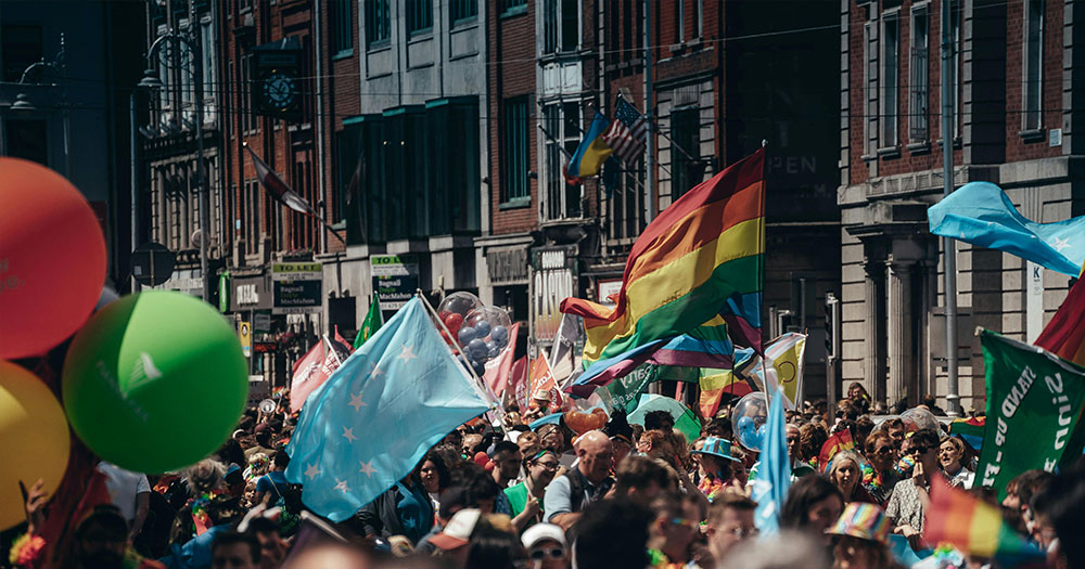 This article is about Ireland's LGBTQ+ history. The image shows a pride parade, a huge crowd with pride and other flags in the background, big colourful balloons.