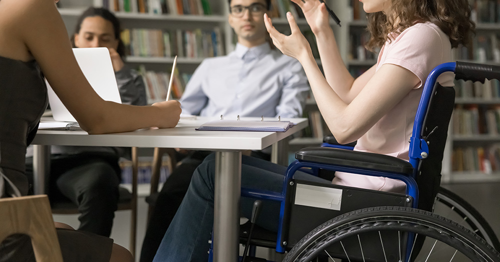 An image of students with disabilities sitting around a desk. The person most in focus uses a wheelchair and is talking to their peers.