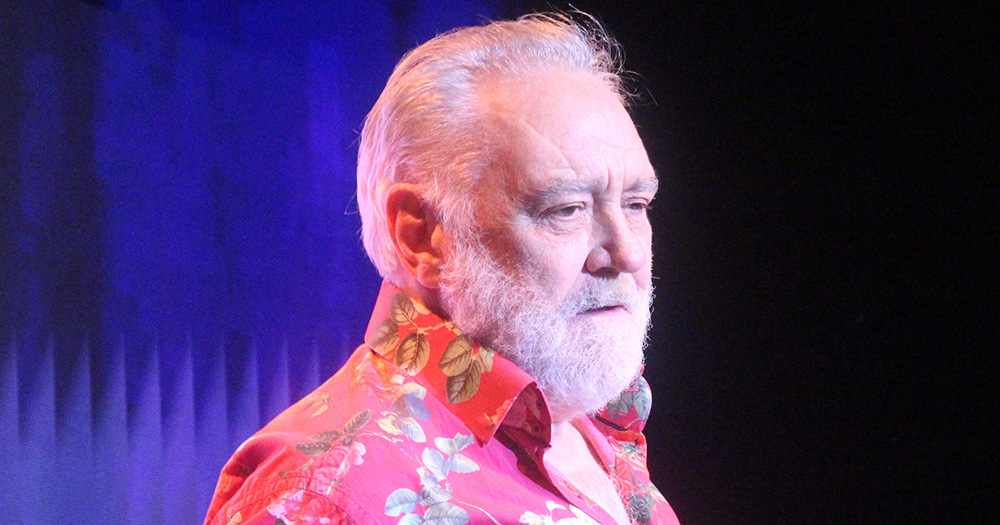 An image of Tony Slattery on stage. He wears a red floral shirt.