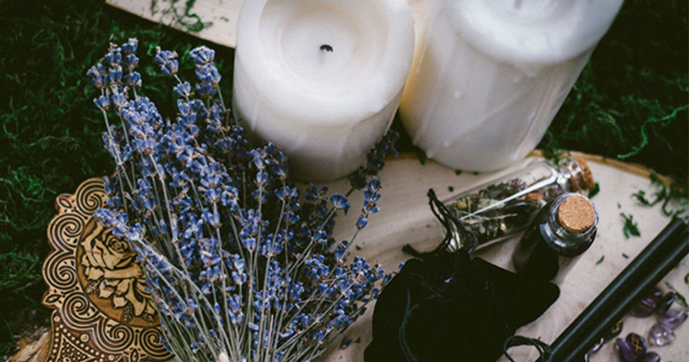 This article is about queer women and witchcraft. Two white burnt pillar candles, a bunch of lavender, a gold mirror, purple crystal runes and a black bag, two black sticks and two corked bottles with dried flowers.