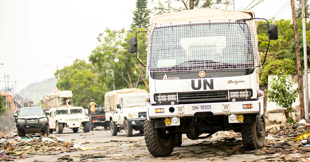 This article is about queer inclusive peacebuilding. The image shows a UN vehicle on debris-littered road.