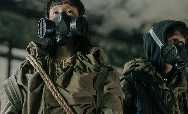 Two people in half face respirators wearing big jackets in a torn down building.