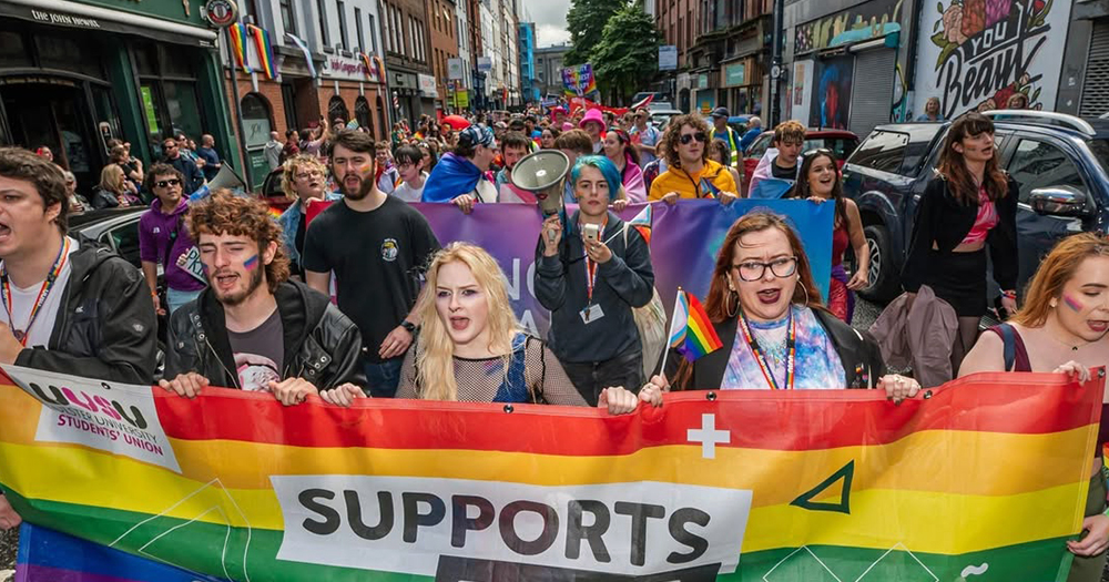 Photo of Belfast Pride, who has recently announced a ban on political parties who voted in favour of banning puberty blockers, with people marching and carrying a Pride flag banner.