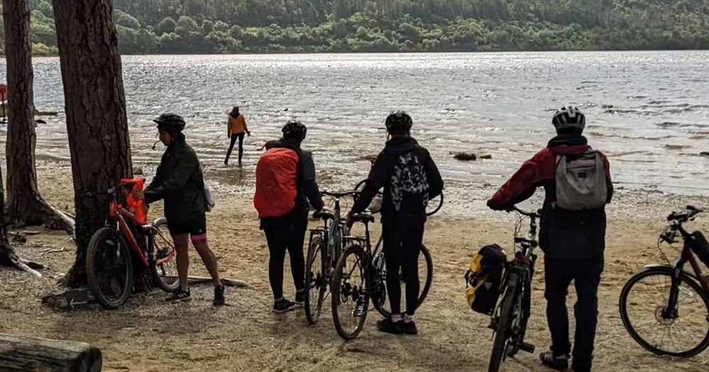 An LGBTQ+ cycling standing beside their bikes on a beach beside a large lake. There are large trees beside them in the sand