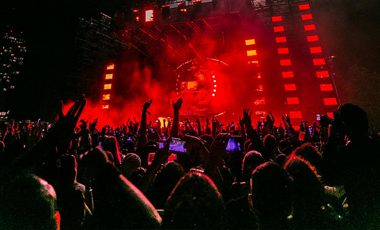 This article is about LGBTQ+ festivals. A large crowd of people surrounding a stage. Smokey with red lights from the stage.