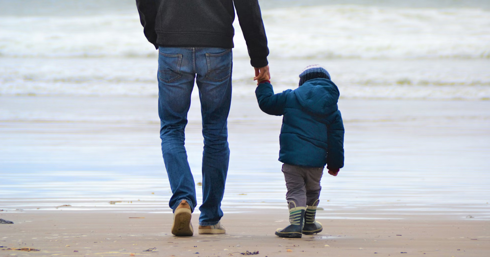 This article is about becoming a foster carer in Ireland. In the photo, a man holding a kid's hand as they walk together towards the sea on a beach.