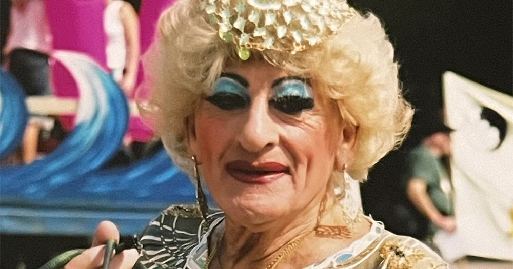 Drag queen Maisie Trollette in full drag, wearing a blonde curly wig and smiling softly at the camera.