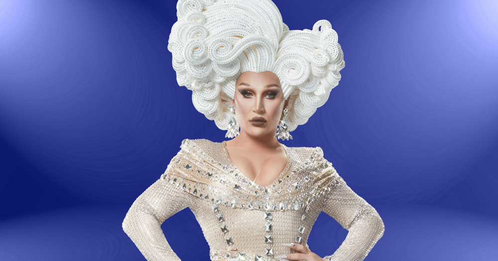 Photo of late drag artist The Vivienne wearing a white dress and wig and posing in front of a blue background.