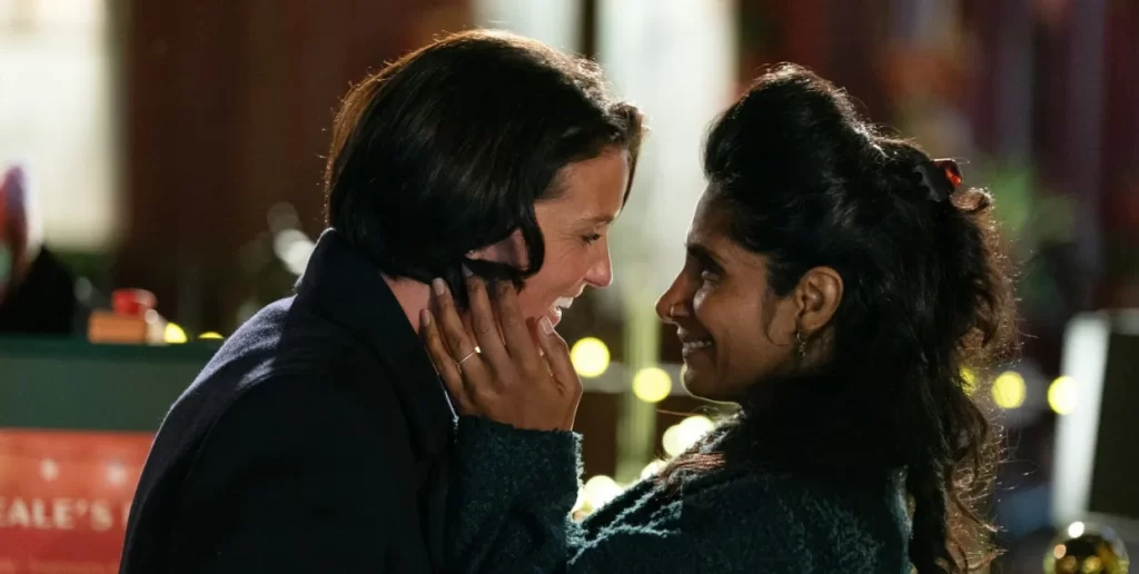 Suki and Eve embrace each other and smile at each other. There are wooded windows and small lights behind them.