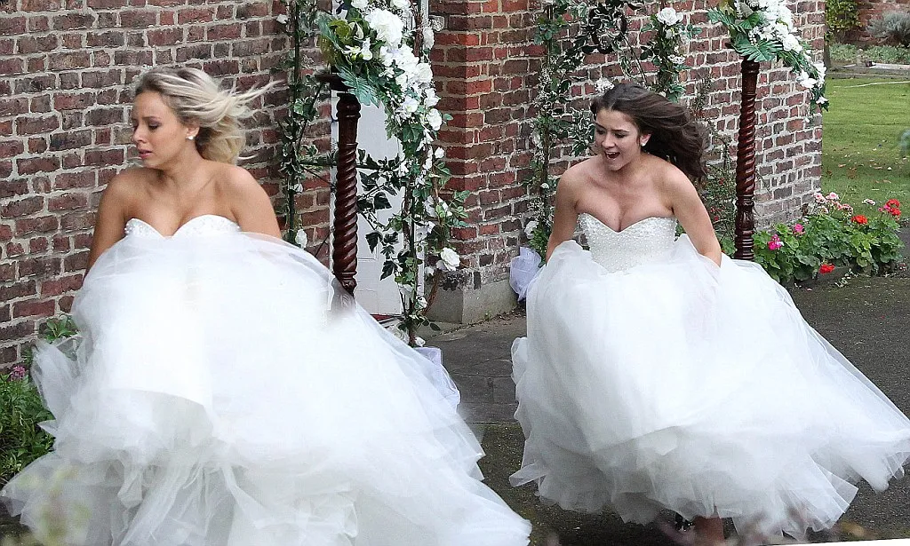 Photo shows Sophie and Sian running beside a red brick building while wearing wedding dresses
