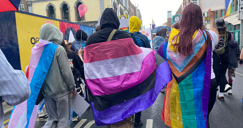The back of three people at Limerick Pride 2024. They each have a different Pride flag draped around their shoulders.
