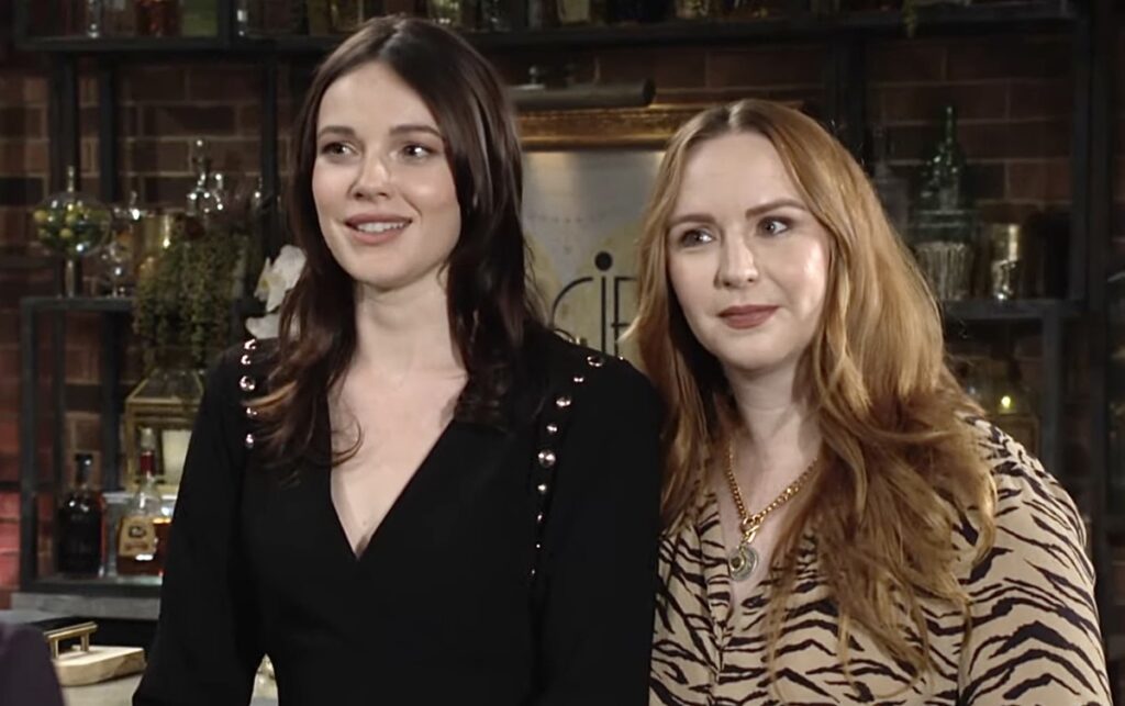 Mariah and Tessa stand beside each other, wearing a black shirt and s beige and black patterned shirt. They both seem pleased, and have lots of glass decorations behind them