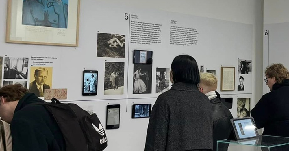 Photo from Poland's LGBTQ+ museum, showing pictures and letters on a white wall. There are a few people standing in front, looking at it.