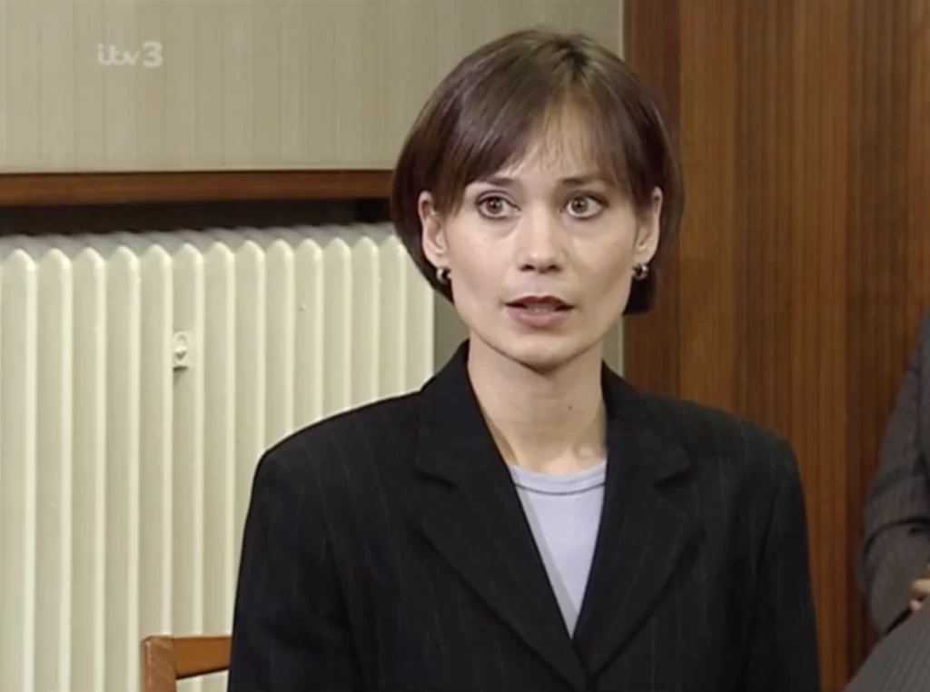 Photo shows Zoe wearing a black blazer and white top. She has a short haircut, and there is a radiator and wooden furnishing behind her.