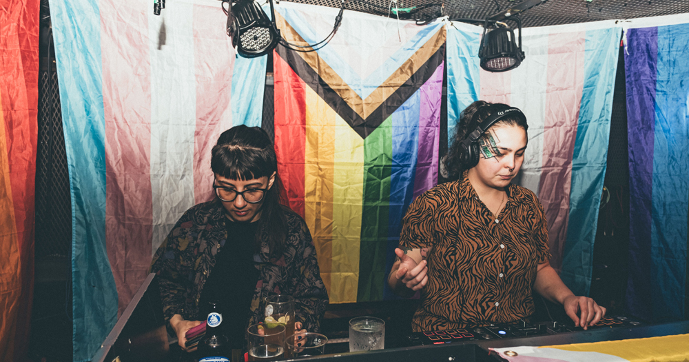 This article is about a partnership between Mother and Barefoot Wine. In the photo, two DJs in front of a progress Pride flag.