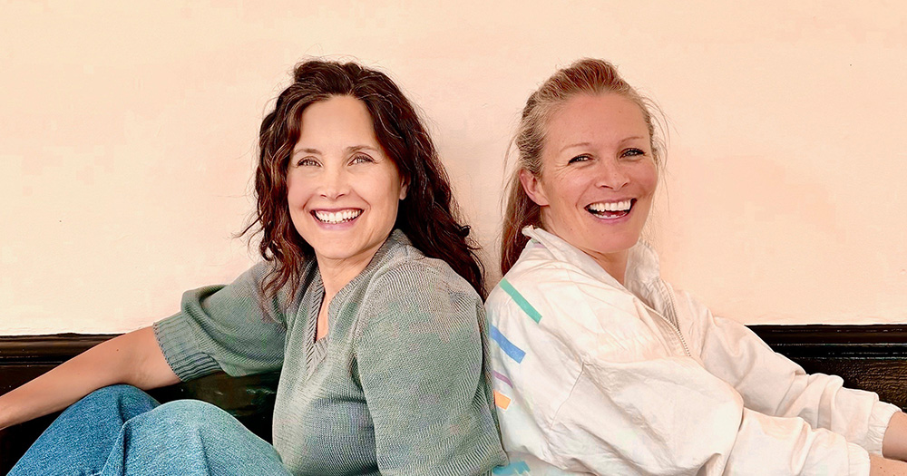 Podcast co-hosts The L Word's Rachel Shelley and former Irish LGBTQ+ hockey player Nikki Symmons sit back to back smiling.