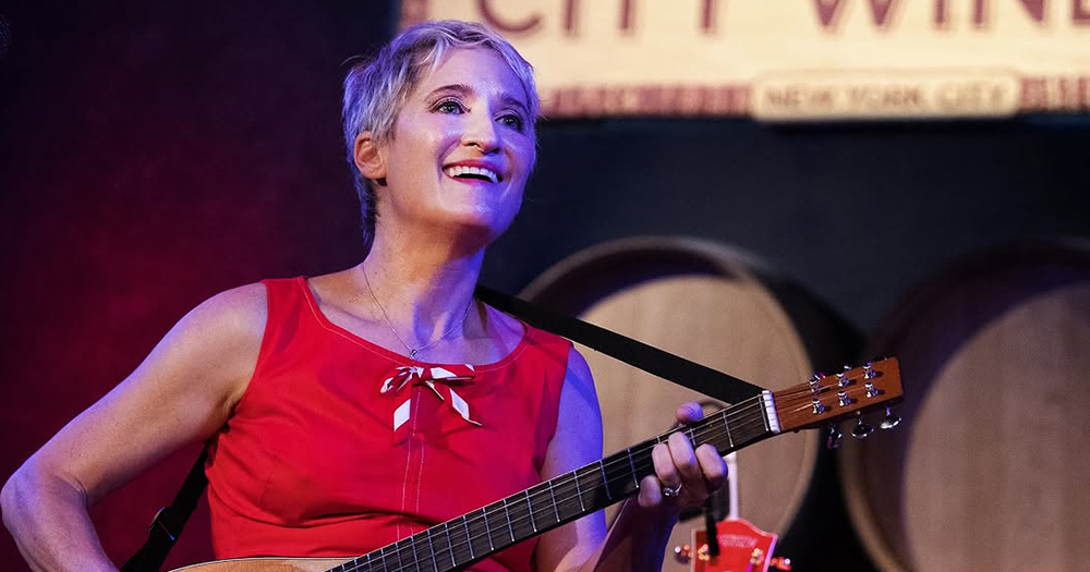 Singer Jill Sobule playing the guitar on a stage and smiling.