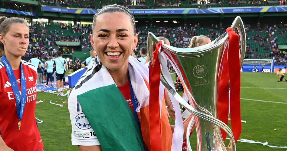 Katie McCabe lifting the Champions League cup, smiling at the camera and wearing an Irish flag.