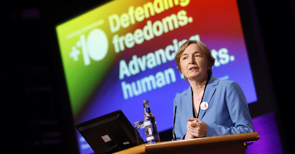 CEO of LGBT Ireland Paula Fagan speaking at a Dublin conference marking 10 years of marriage equality in Ireland. Fagan is speaking into a microphone on a stage with a screen in the background that reads "Defending freedoms, advancing human rights".