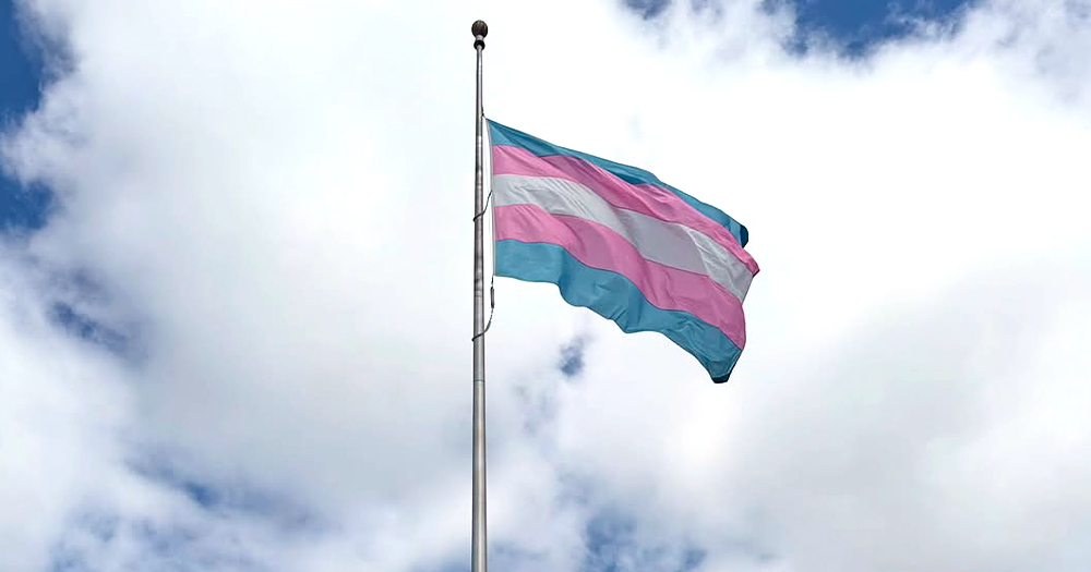 This article is about trans rights regressing in Europe. In the photo, a trans flag flying in a cloudy sky.