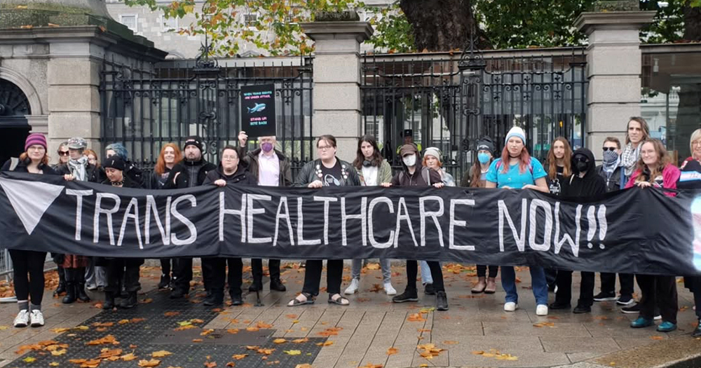 Activist gathered at a protest holding a banner that reads "Trans healthcare now".