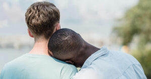 The back of a gay couple travelling together. They are photographed outside, with the man on the right resting his head on his boyfriend's shoulder.