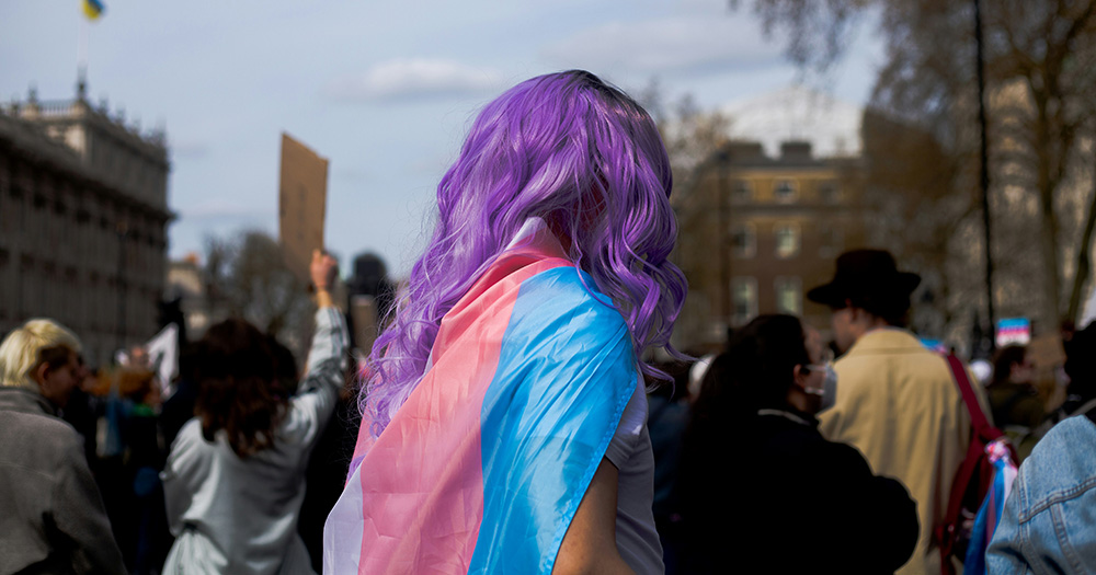 This article is about gender law reforms in Western Australia. The image shows the back of a person with purple hair with a trans flag around their shoulders.