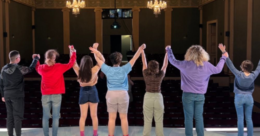 For the theatre week of Belong To's summer project, a group of queer youth standing hand in hand with their arms raised, poised to take a bow on stage.