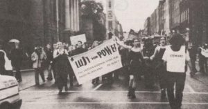Photo of first Dublin Dyke March with people marching and carrying a sign that reads "Lust for Power".