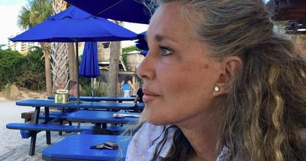 Profile shot of Caroline Cossey sitting outside at a beach area surrounded by blue picnic tables with blue umbrellas.
