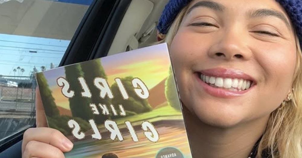 Hayley Kiyoko in a car holding a copy of her book, Girls Like Girls
