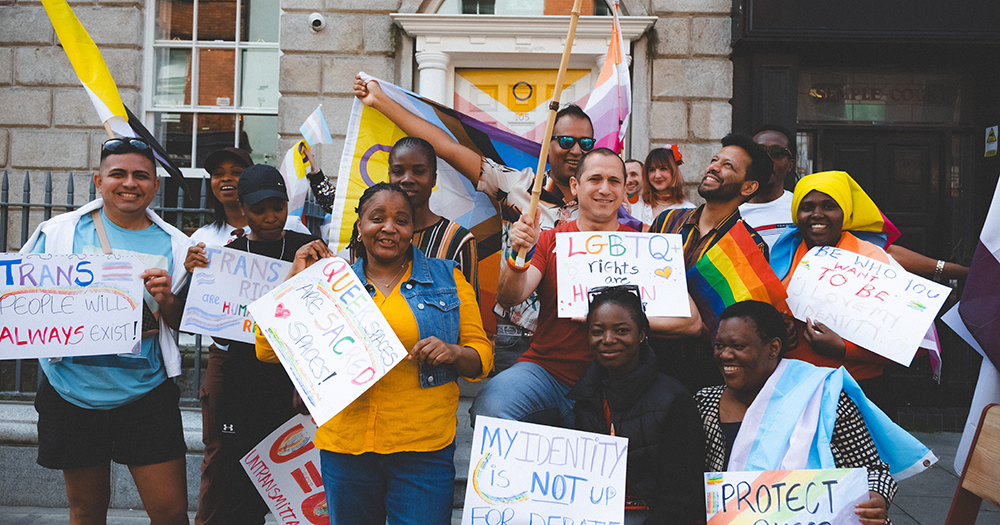 A group of people hold Pride flags and banners outside Outhouse LGBTQ+ Centre.