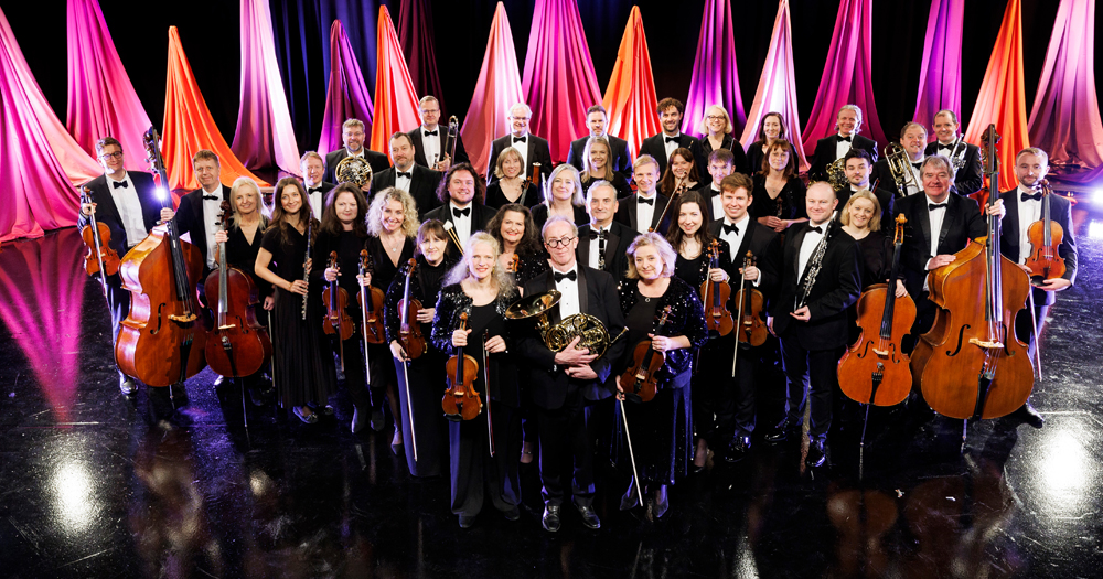 Members of the RTÉ Concert Orchestra posing on stage dressed in all black with pink drapes in the background.
