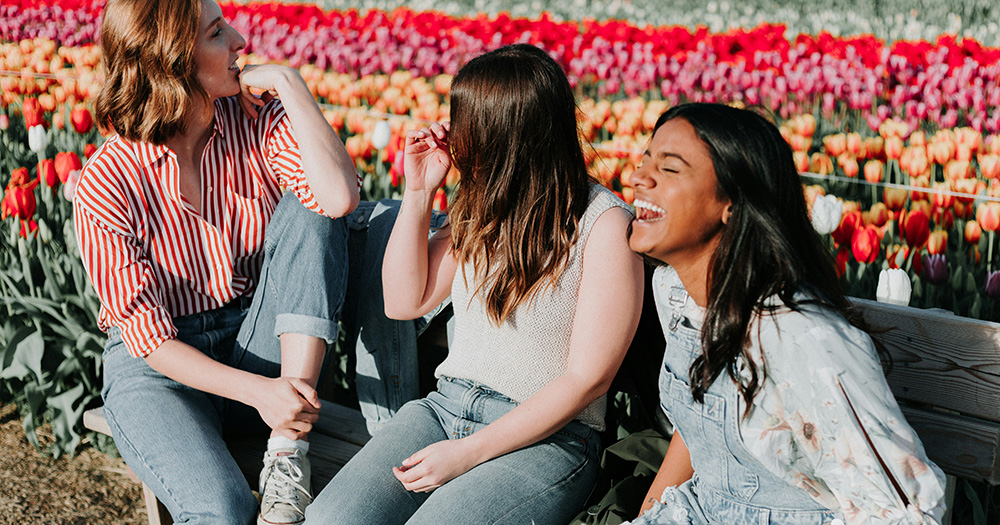 This article is about staying sober at Pride and beyond. The image shows three women laughing with rows of colourful flowers in the background.